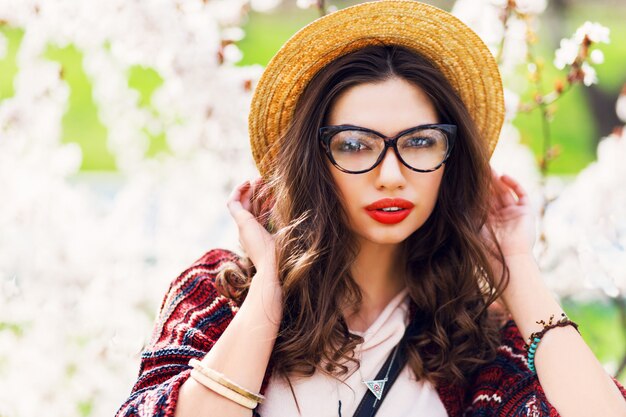 Femme incroyable avec maquillage lumineux, yeux bleus, lunettes, chapeau de paille posant dans un parc printanier ensoleillé près de l'arbre à fleurs