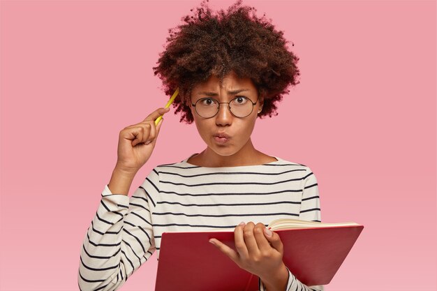 Femme indécise confuse à la peau noire, cheveux bouclés, se gratte la tête avec un crayon, vêtue d'un pull rayé, lèvres de bourses