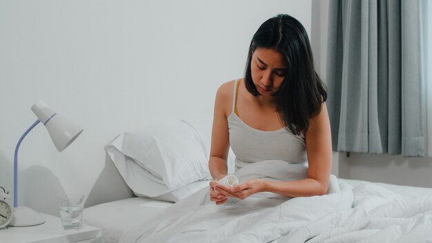 Femme indienne malade et en mauvaise santé souffre d'insomnie. Asiatique jeune femme prenant des médicaments antidouleur pour soulager les maux de tête et boire un verre d'eau assis sur son lit dans sa chambre à la maison le matin.