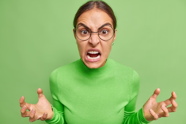 Photo gratuite une femme indignée en colère regarde avec des gestes de rage et fronce les sourcils, le visage irrité par quelque chose qui porte des lunettes rondes pose décontractée à col roulé contre un mur vert vif