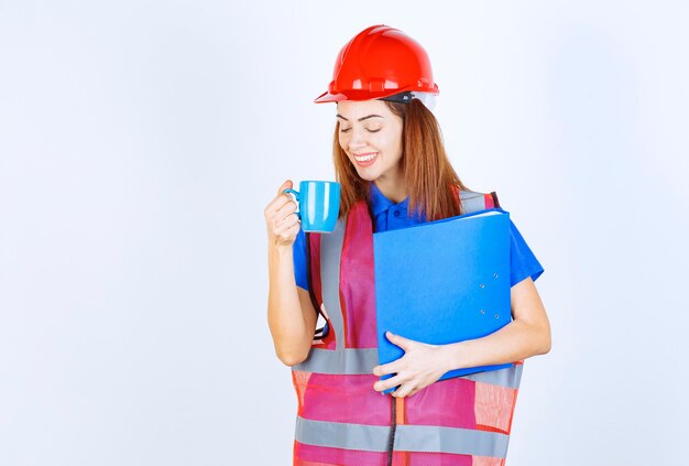 Femme ingénieur en casque rouge tenant un dossier bleu et buvant une tasse de boisson.