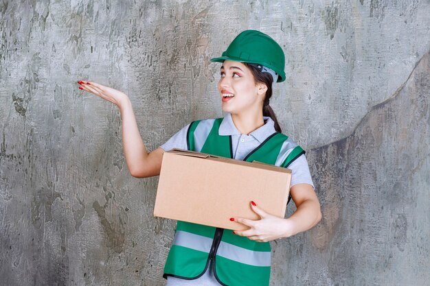 Femme ingénieur en casque vert tenant une boîte en carton et pointant vers quelqu'un autour