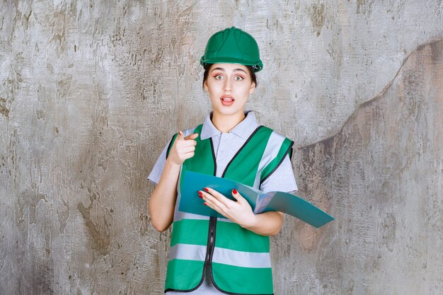 Femme ingénieur en casque vert tenant un dossier bleu et pointant vers quelqu'un autour