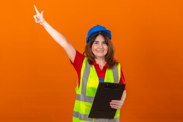 Femme ingénieur en gilet de construction et casque de sécurité tenant le presse-papiers dans la main à la confiance avec le sourire sur le visage heureux pointant vers quelque chose avec le doigt debout sur backgro orange isolé