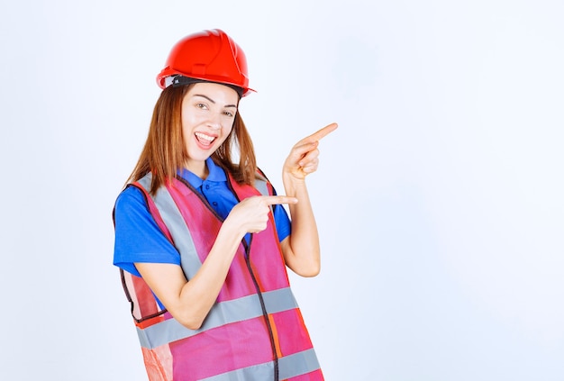 Femme ingénieur en uniforme et casque rouge pointant vers quelqu'un sur la droite.