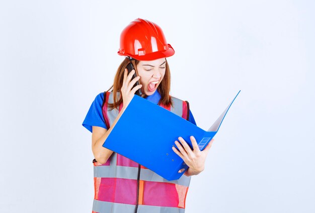 Femme ingénieur en uniforme et casque rouge tenant un dossier bleu et criant au téléphone.