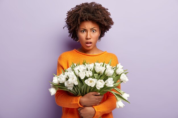 Une femme insatisfaite indignée a l'air en colère, tient des fleurs blanches, porte un pull orange, des mannequins sur un mur violet, exprime des émotions négatives, entend de mauvaises nouvelles. Femme aux tulipes