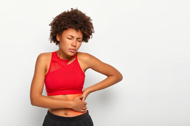 Une femme insatisfaite tient la hanche douloureuse, a une inflammation des reins, touche l'emplacement de la douleur près des côtes marquées d'un point rouge, porte un soutien-gorge de sport