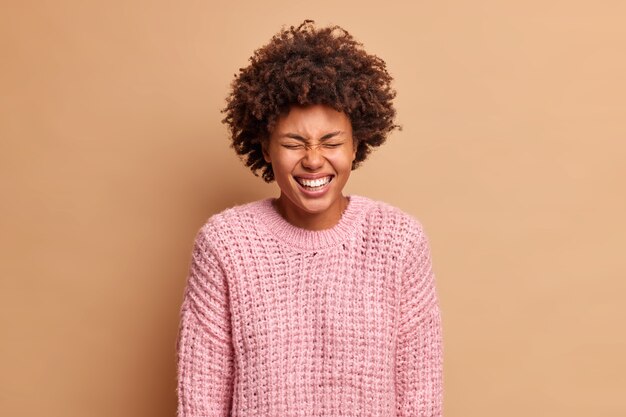 Une femme insouciante amusée ferme les yeux et sourit exprime largement des émotions pures parle à un ami drôle porte un chandail tricoté chaud pose contre le mur beige rit fort