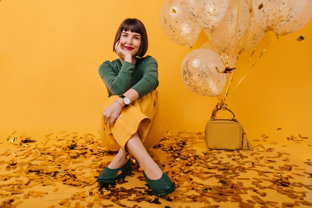 Femme insouciante avec coupe de cheveux courte assise avec un sourire doux. Portrait intérieur d'une fille séduisante entourée de confettis et de ballons de fête.