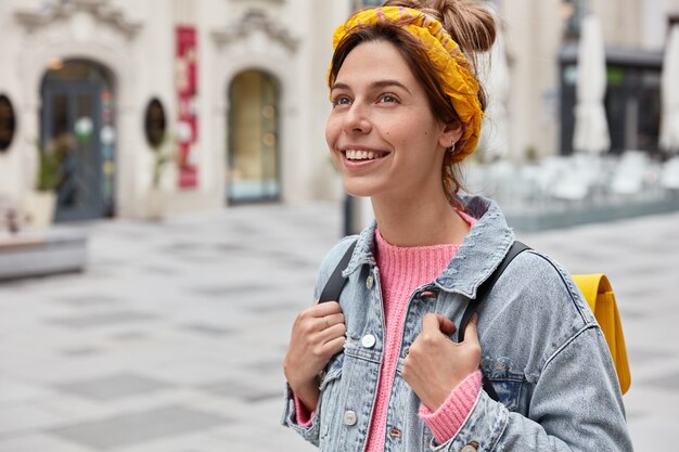 Femme insouciante joyeuse habillée en tenue à la mode, a un sourire charmant sur le visage, aime les loisirs, porte un petit sac à dos