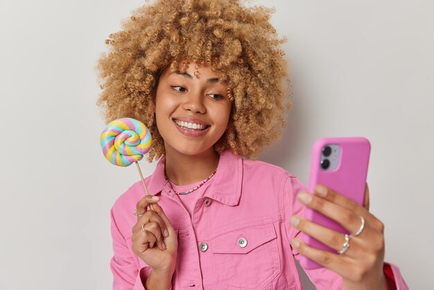 Une femme insouciante positive aux cheveux bouclés tient des bonbons multicolores prend un selfie sur l'appareil photo d'un smartphone vêtue d'une veste rose à la mode isolée sur fond blanc