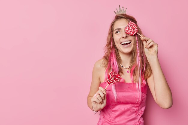 Une femme insouciante positive couvre les yeux avec des sourires de sucette en forme de coeur porte avec joie une petite couronne et une robe regarde loin isolée sur fond rose avec un espace de copie pour votre contenu promotionnel