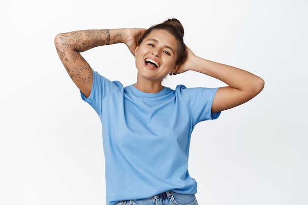 Femme insouciante riant et se reposant, tenant les mains derrière la tête dans une pose insouciante, souriante heureuse, regardant quelque chose, debout sur fond blanc