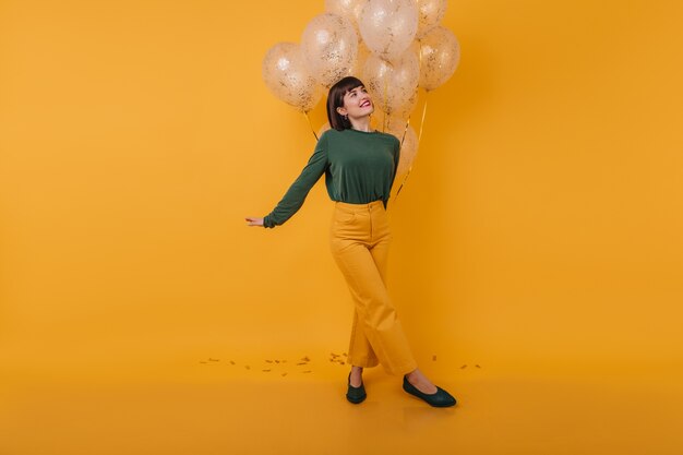 Femme inspirée avec une coupe de cheveux à la mode en détournant les yeux tout en posant avec des ballons de fête. Photo intérieure d'une incroyable dame caucasienne en tenue vintage s'amusant après la fête.