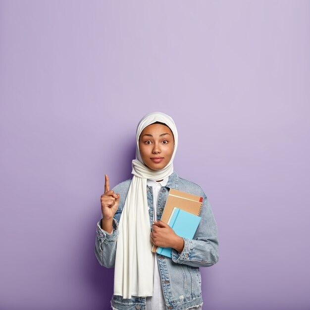 Une femme intrigante surprise en couvre-chef pointe vers le haut et regarde avec intérêt, montre un espace vide au-dessus pour votre publicité ou information, porte un journal et un cahier à spirale. Religion musulmane