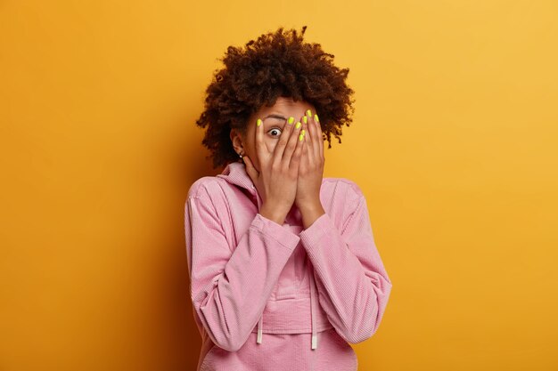 Une femme intriguée et curieuse à la peau sombre jette un coup d'œil à travers les doigts, se couvre le visage avec des paumes, se cache, a peur de quelque chose, a les yeux écarquillés, vêtue d'un sweat-shirt décontracté, pose à l'intérieur sur un mur jaune