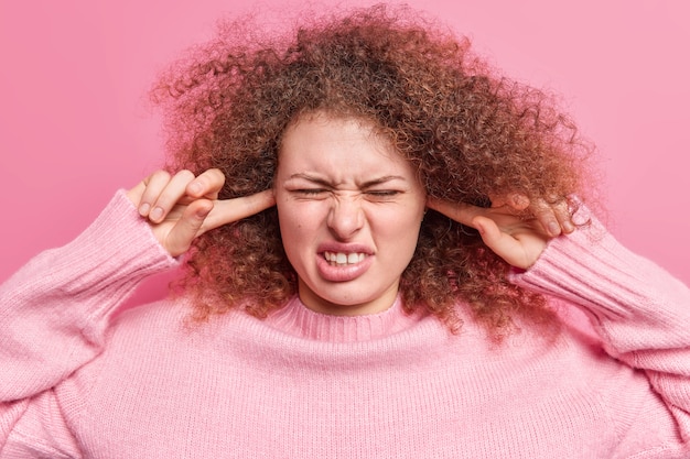 Une femme irritée aux cheveux bouclés se bouche les oreilles pour éviter les bruits forts ne peut pas supporter un son agaçant ne peut pas se concentrer dans un endroit bruyant et bondé porte un pull décontracté se sent mal à l'aise isolé sur un mur rose