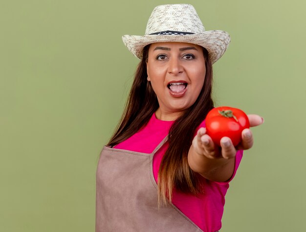 Femme de jardinier d'âge moyen en tablier et chapeau offrant des tomates fraîches regardant la caméra en souriant debout sur fond clair