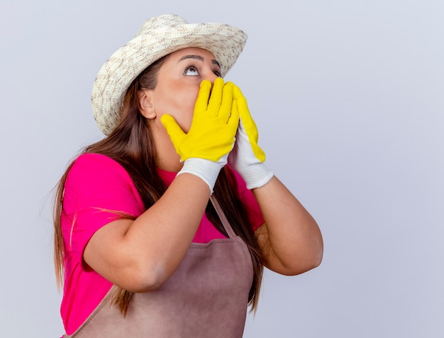 Femme de jardinier d'âge moyen en tablier et chapeau portant des gants en caoutchouc à la recherche d'être choqué debout sur fond blanc