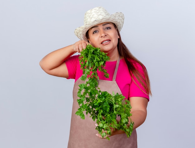 Femme de jardinier d'âge moyen en tablier et chapeau tenant des herbes fraîches regardant la caméra en souriant joyeusement debout sur fond blanc