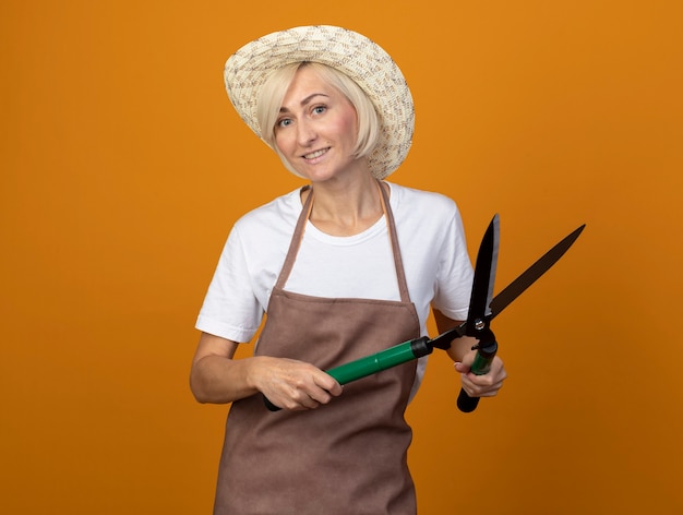 Photo gratuite femme jardinière blonde d'âge moyen souriante en uniforme portant un chapeau tenant des cisailles à haie