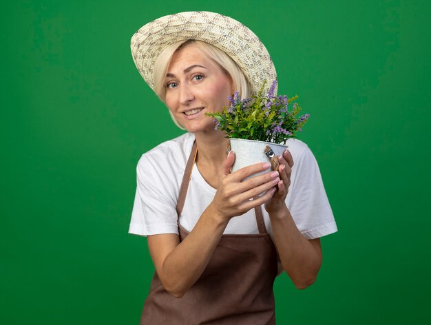 Femme jardinière blonde d'âge moyen souriante en uniforme portant un chapeau tenant un pot de fleurs