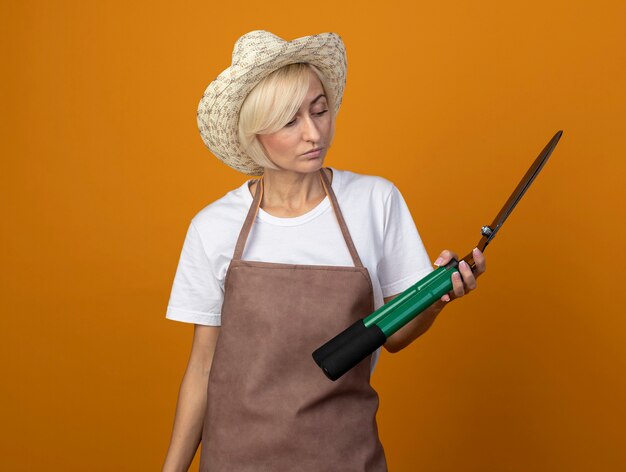 Photo gratuite femme jardinière blonde d'âge moyen en uniforme portant un chapeau tenant et regardant des cisailles à haies isolées sur un mur orange avec espace de copie