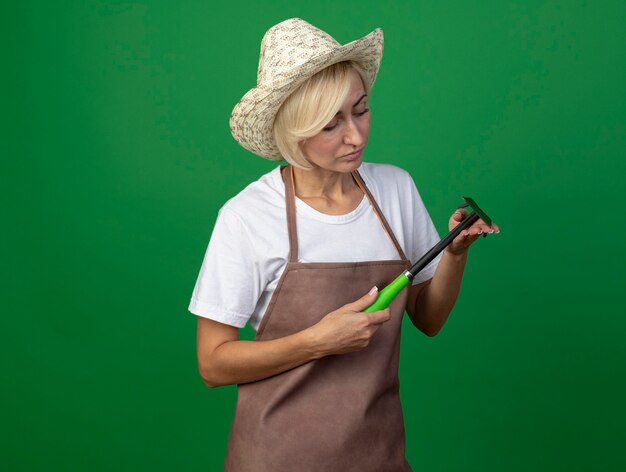 Femme jardinière blonde d'âge moyen en uniforme portant un chapeau tenant et regardant un râteau-houe
