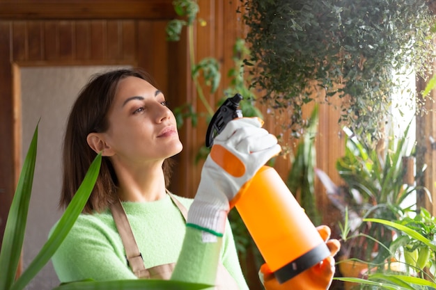 Femme jardinière à la maison en tablier et gants avec des plantes en croissance sur le balcon de la maison arrosant à l'aide d'un spray