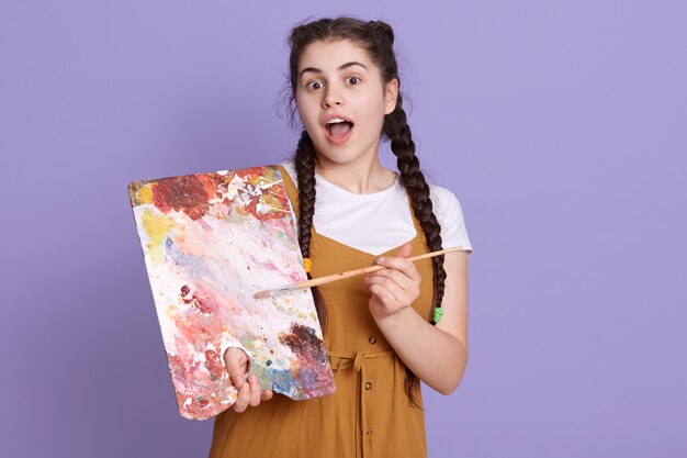 Femme jeune artiste brune avec des nattes tenant un pinceau et une palette de peintre sur un mur lilas, posant avec un visage surprise, debout avec la bouche ouverte.