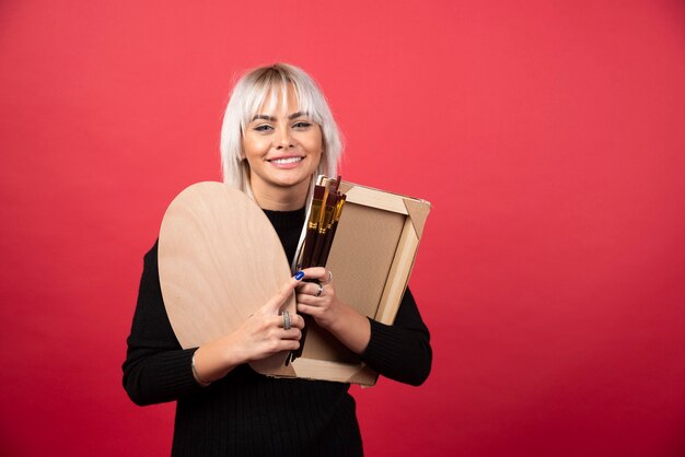 Femme jeune artiste tenant des fournitures d'art sur un mur rouge.