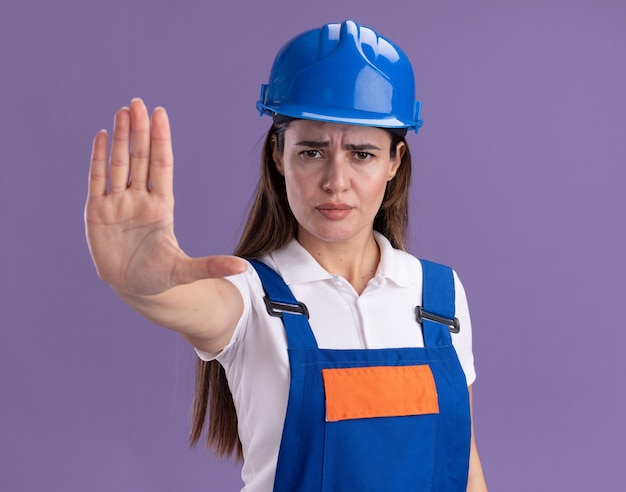 Femme jeune constructeur strict en uniforme montrant le geste d'arrêt isolé sur mur violet