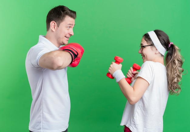 Femme jeune couple sportif avec des haltères en regardant son petit ami avec des gants de boxe souriant joyeusement debout sur mur vert