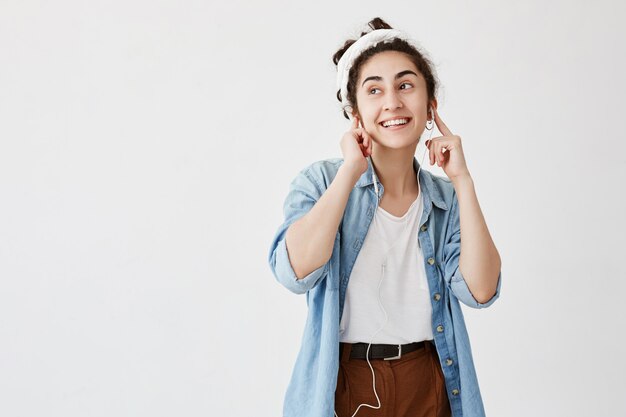 Femme ou jeune heureuse et expressive aux cheveux foncés et bouclés en chignon, écoute de la musique dans les écouteurs, aime les mélodies agréables, a la bonne humeur. Gens, loisirs, style de vie, beauté, concept de bonheur