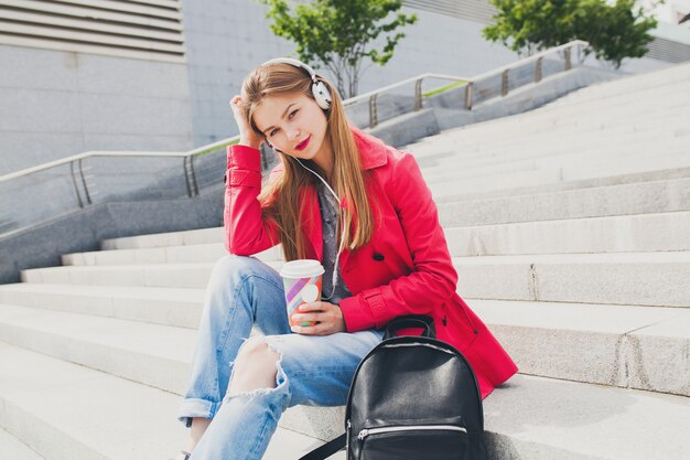 Femme jeune hipster en manteau rose, jeans assis dans la rue avec sac à dos et café en écoutant de la musique au casque, tendance de style printemps urbain grande ville