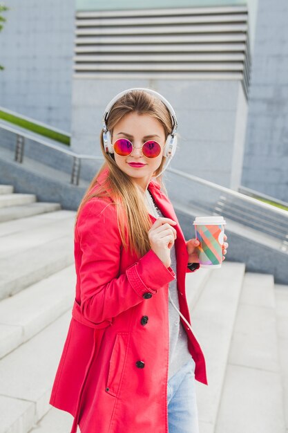 Femme jeune hipster en manteau rose, jeans dans la rue avec du café en écoutant de la musique au casque