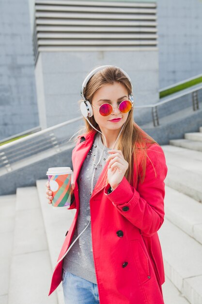 Femme jeune hipster en manteau rose, jeans dans la rue avec du café, écouter de la musique au casque, porter des lunettes de soleil