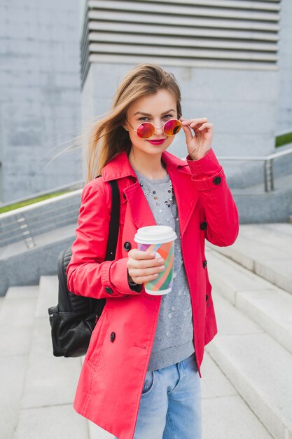 Femme jeune hipster en manteau rose, jeans dans la rue avec sac à dos et café, écouter de la musique au casque, porter des lunettes de soleil