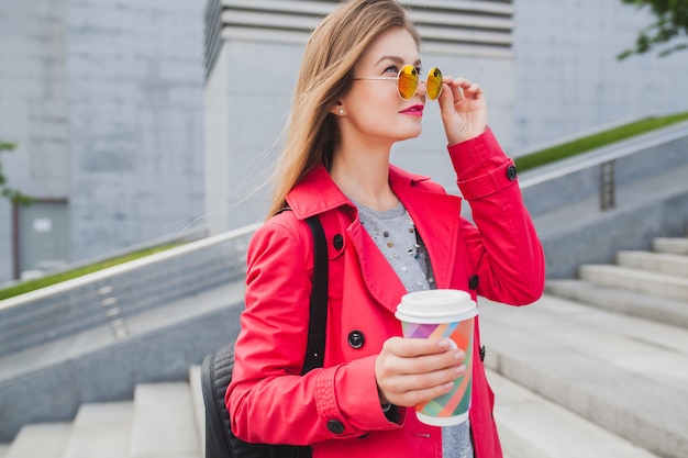 Femme jeune hipster en manteau rose, jeans dans la rue avec sac à dos et café, écouter de la musique au casque, porter des lunettes de soleil