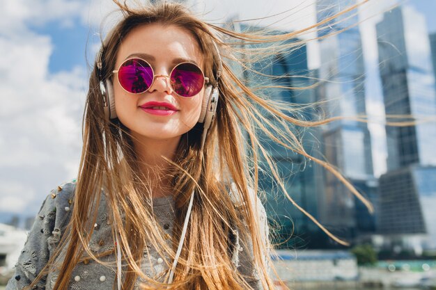 Femme jeune hipster s'amuser dans la rue, écouter de la musique sur des écouteurs, portant des lunettes de soleil roses