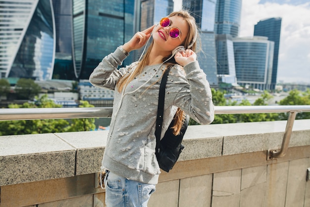 Femme jeune hipster s'amuser dans la rue, écouter de la musique sur des écouteurs, portant des lunettes de soleil roses