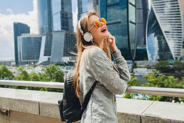 Femme jeune hipster s'amuser dans la rue, écouter de la musique sur des écouteurs, porter des lunettes de soleil roses, style urbain printemps été