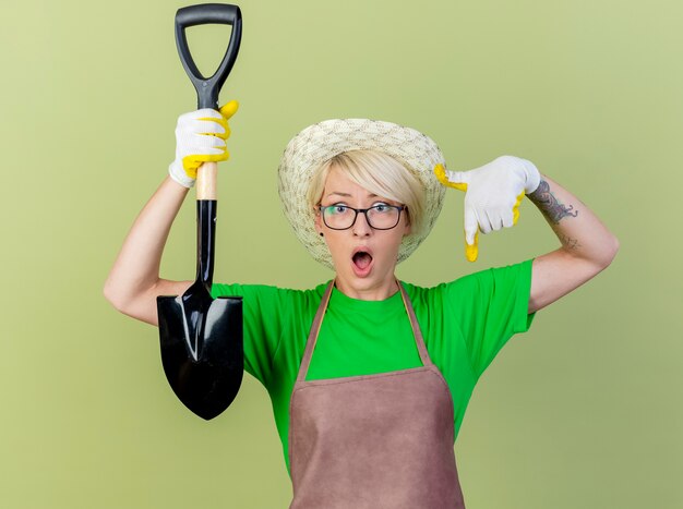 Femme jeune jardinier aux cheveux courts en tablier et chapeau portant des gants en caoutchouc tenant une pelle pointant avec l'index à la surprise debout sur fond clair