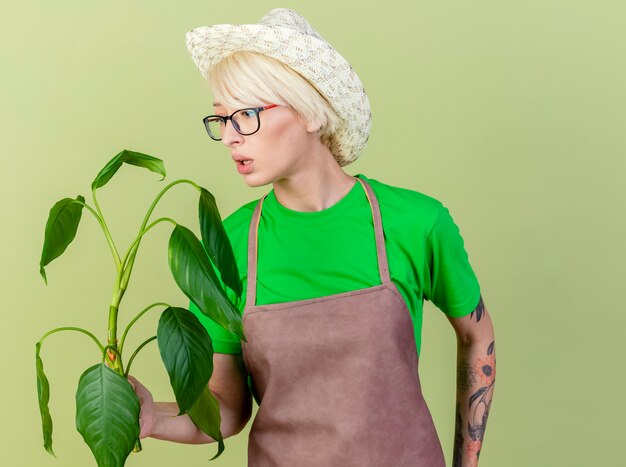 Femme jeune jardinier aux cheveux courts en tablier et chapeau tenant une plante