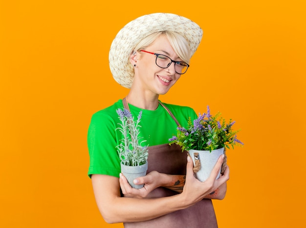 Femme jeune jardinier aux cheveux courts en tablier et chapeau tenant des plantes en pot souriant joyeusement heureux et positif debout sur fond orange