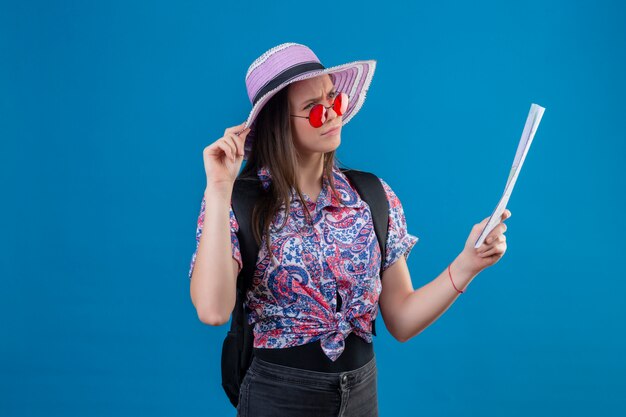 Femme jeune voyageur en chapeau d'été portant des lunettes de soleil rouges tenant la carte à côté avec une expression pensive pensant avoir des doutes sur le mur bleu