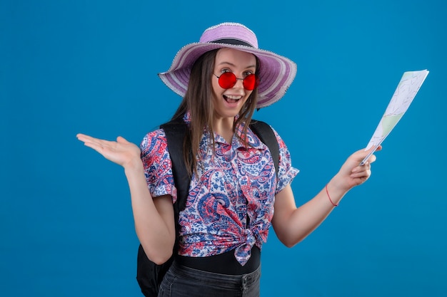 Femme jeune voyageur en chapeau d'été portant des lunettes de soleil rouges tenant la carte à la position positive et heureuse avec le bras levé sur fond bleu