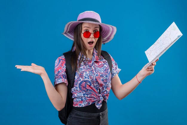 Femme jeune voyageur en chapeau d'été portant des lunettes de soleil rouges tenant la carte regardant la caméra étonné et surpris avec une large bouche d'ope debout sur fond bleu