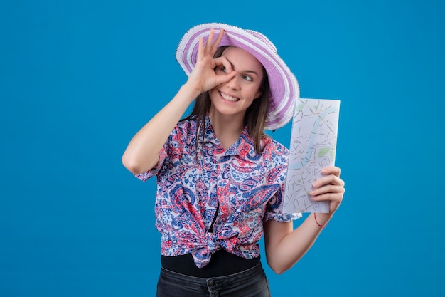 Femme jeune voyageur en chapeau d'été tenant la carte positive et heureuse de faire signe ok regardant à travers ce signe debout sur fond bleu